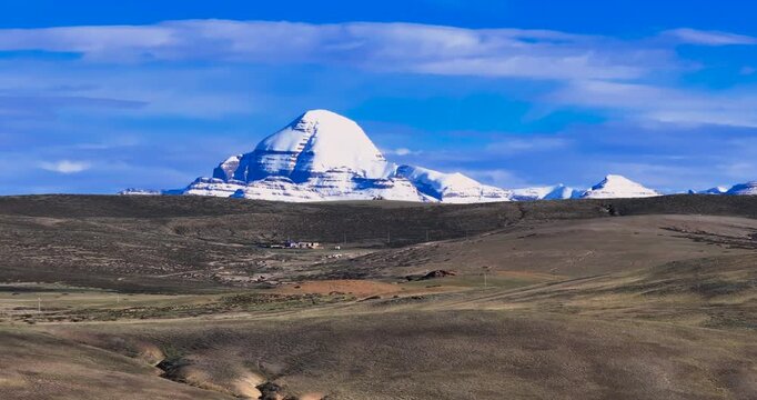 A lateral drone glide shot focuses on the Mount Kailash mountain range in Tibet, showcasing majestic snow-covered Himalayan peaks dominating the frame above arid terrain and open land.