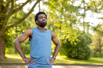 Fototapeta premium Man standing waist-up in a sunlit green park, eyes closed and hands on hips, taking a deep breath to recover from a run, calm and focused on fitness outdoors