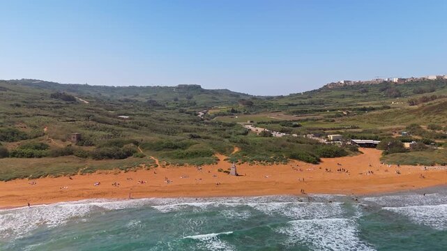 Ramla Beach on Gozo, Malta features distinctive red sand and turquoise Mediterranean waters bordered by rural countryside and fields, seen from a drone perspective on a sunny day.