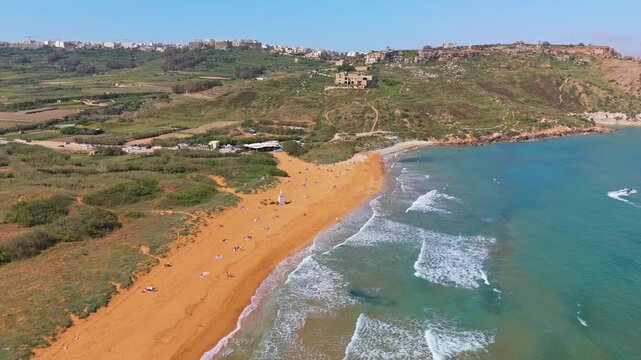 Ramla Beach on Gozo features distinctive reddish sand bordered by turquoise Mediterranean waters, green hillsides, and coastal cliffs, viewed in a panoramic aerial perspective on a sunny day.