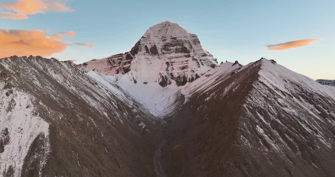 Drone footage captures the iconic, snow-covered peak of Mount Kailash in Tibet rising between steep mountain ridges beneath a clear evening sky with scattered clouds in low-angle light