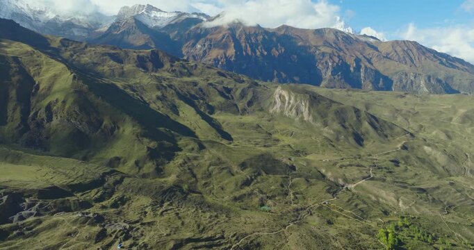 A drone glide shot tracks over green hills and sloping terrain in Mustang, Nepal, revealing a layered Himalayan mountain range in the background under natural daylight.