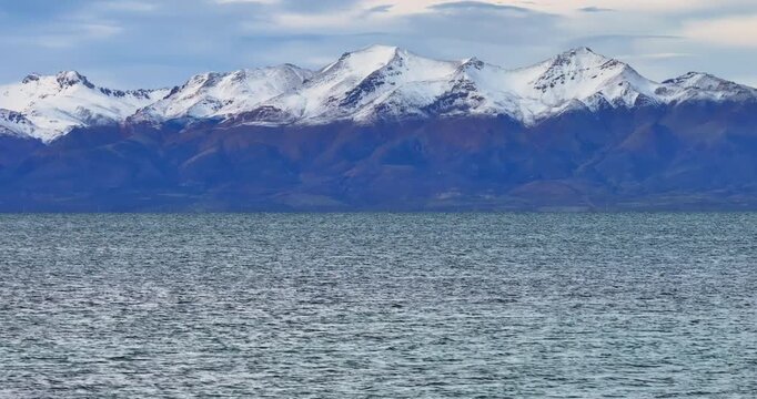 A steady drone shot moves smoothly over the wide expanse of Lake Manasarovar, capturing the rippling blue water in the foreground and the snow-capped Gurla Mandhata mountain range under blue sky.