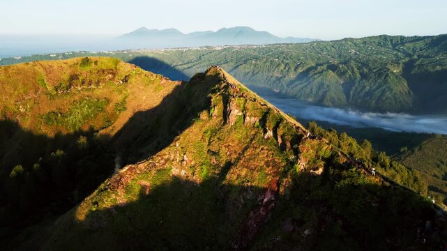 Hikers trekking along narrow volcanic ridge at sunrise, exposed ridge trail with caldera views and rugged mountain terrain in Bali Indonesia landscape, rising drone footage