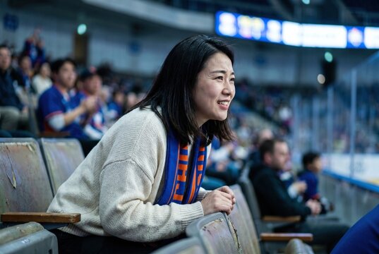 Excited female sports fan watching a game from arena seats, leaning forward and smiling while wearing a blue and orange team scarf