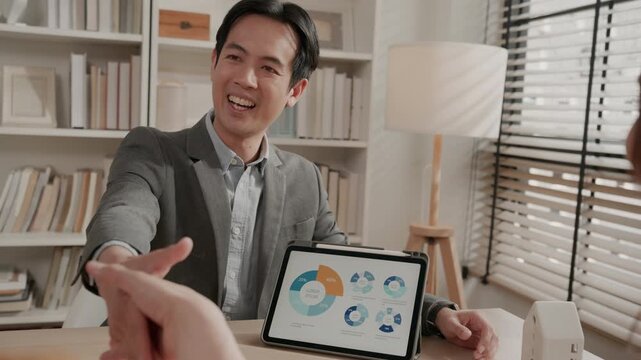 Male financial advisor presenting savings or investment strategies to a young Asian couple. Advisor smiling and shaking hands with client to close a deal. Wealth management, real estate investment.