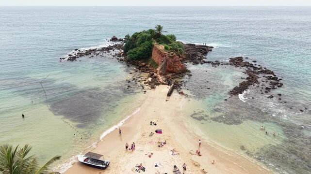 Aerial view of Parrot Rock island connected by a sandbar to the beach with people and palm trees Mirissa, Southern Province, Sri Lanka.