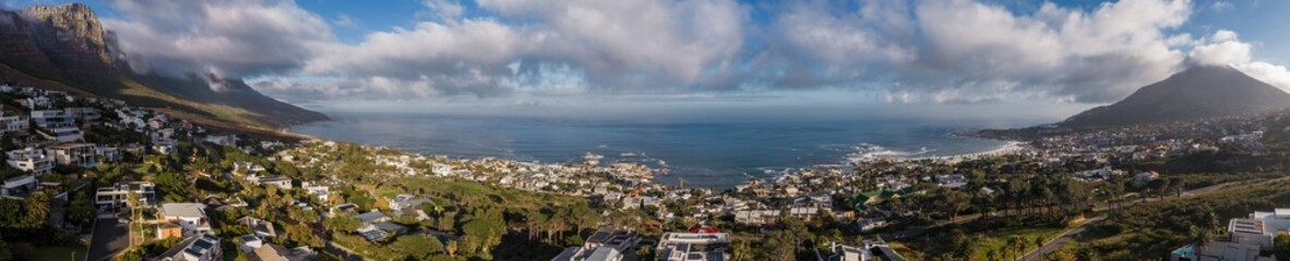 Fototapeta premium Aerial view of Cape Town Atlantic Seaboard shows suburbs on green hills, Twelve Apostles by Camps Bay, Lion's Head at right, calm steel blue ocean, late afternoon light.