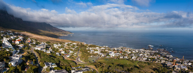Aerial panorama of Cape Town's Atlantic Seaboard shows Twelve Apostles with low clouds, Camps Bay and Clifton curving to the right, white houses, coves, and calm ocean. © Aerial Film Studio