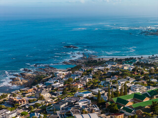 Aerial view of Cape Town Atlantic Seaboard shows Camps Bay and Clifton with whitewashed homes, green roofs, rocky shore, turquoise coves, boulders, and tidal pools. © Aerial Film Studio