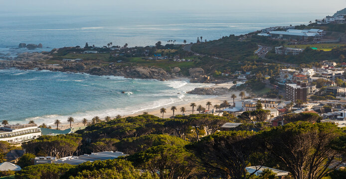 Elevated coastal view of Camps Bay, Cape Town, with palms, gentle surf, low rise hotels, granite outcrops, fynbos slopes, and winding coastal roads in late afternoon sun.