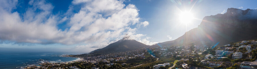 Wide panoramic view of Cape Town Atlantic seaboard, sunlit clouds drift, Lion's Head rises, Twelve Apostles at right, whitewashed houses step to ocean, bright daytime. © Aerial Film Studio
