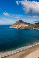 Obraz premium Aerial view of Hout Bay in Cape Town shows sandy beach, turquoise to deep blue water, curving harbor breakwater, rugged mountains, and late afternoon light under clouds.