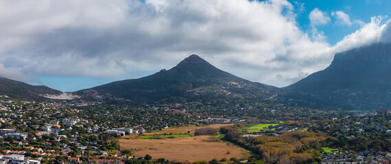 Lion's Head stands centered under drifting clouds, Table Mountain looms right. Cape Town suburbs and winding roads frame a tawny wetland in late afternoon light. © Aerial Film Studio