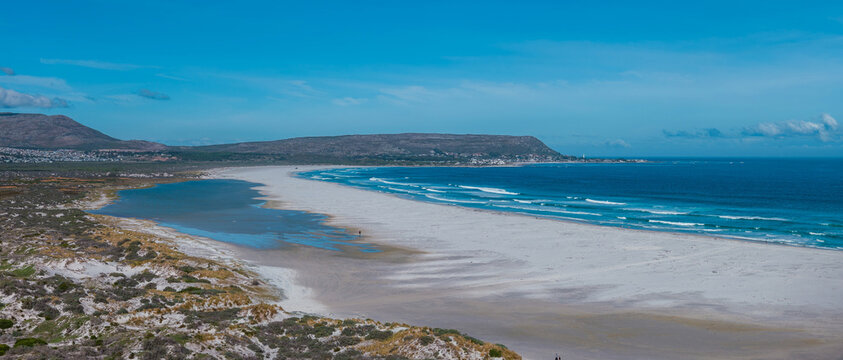 Aerial view shows a wide beach curving by turquoise waters near Cape of Good Hope, with scrubby dunes, fynbos, a flat topped ridge, a lagoon, and few distant figures.