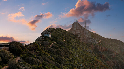 Obraz premium Cape Point in Table Mountain National Park, near Cape Town, shows a lighthouse and tall masts on a rocky summit, zigzag footpaths, fynbos, and sea cliffs at sunset.