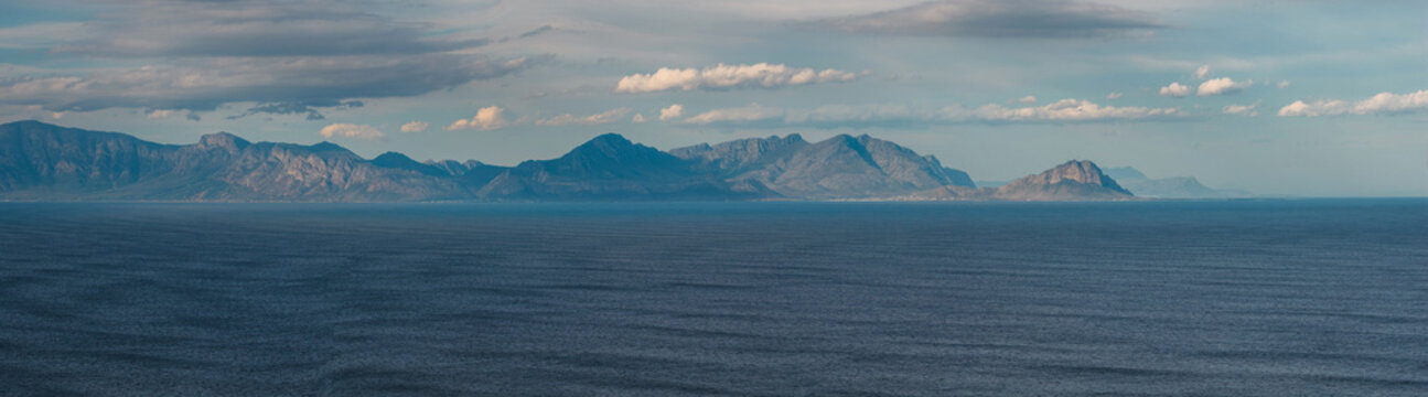 Wide seascape shows steel blue water leading to the Cape Peninsula and layered peaks across False Bay, Cape Town, under soft clouds, cool daylight, and long lines.