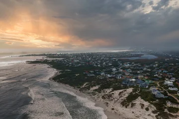 Aerial view of a coastal suburb near Cape Town as Atlantic surf meets a curving sandy beach and dunes, with rooftops by a lagoon and storm clouds glowing at sunset. © Aerial Film Studio