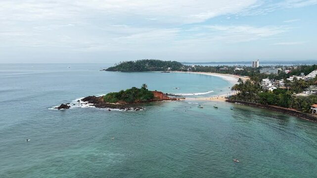 Aerial view of Parrot Rock connected to the beach by a sandbar with turquoise ocean waves and surfers in Mirissa, Southern Province, Sri Lanka.