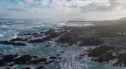 Fototapeta premium Aerial view of the stormy Atlantic near Cape Agulhas in South Africa, with dark rock outcrops, teal sea, whitecaps, low dunes, and sparse plants under a windy, cloud streaked sky.