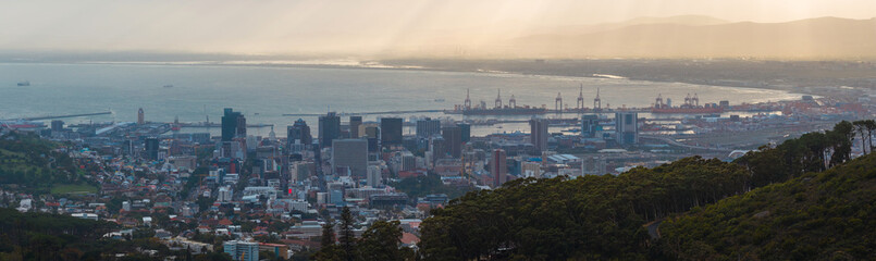 Obraz premium Panoramic elevated view of Cape Town CBD toward Table Bay, Port of Cape Town cranes, anchored vessels, curved shoreline, soft warm light, haze, hills in shadow.
