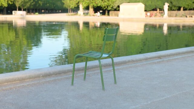 Iconic green metal chair sitting on a gravel path next to a calm reflection pond in a beautiful public park in Paris.