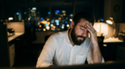 Obraz premium Soft focus blurred photo of Tired man with a beard feeling stressed while working late at his office desk with city lights visible in the background at night