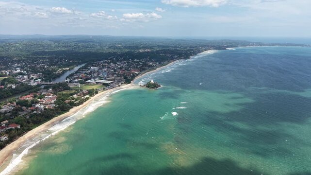 Aerial view of Parewi Duwa Temple on a small island surrounded by turquoise ocean waves and the sandy coastline of Matara, Sri Lanka.