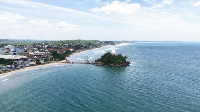 Aerial view of Parewi Duwa Temple on a small island connected by a bridge to the sandy beach and coastal town of Matara, Sri Lanka.