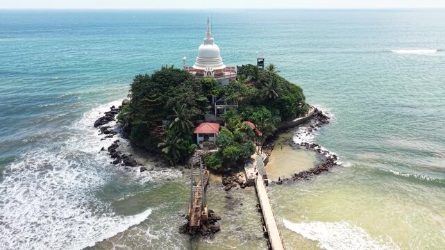 Aerial view of Parewi Duwa Temple, a white Buddhist stupa on a lush green island surrounded by turquoise ocean waves and a bridge, Matara, Sri Lanka.