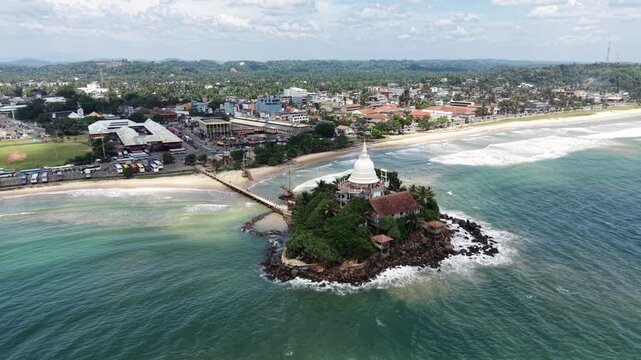 Aerial view of Parewi Duwa Temple on a rocky island connected by a bridge to the coast with city buildings and waves in Matara, Sri Lanka.