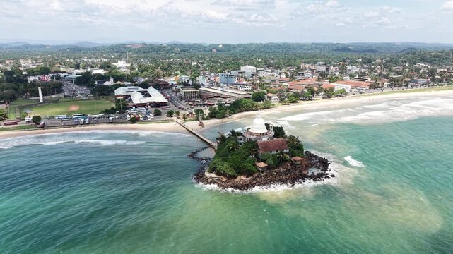 Aerial view of Parewi Duwa Temple on a small island connected by a bridge to the coast with turquoise waves and city skyline in Matara, Sri Lanka.