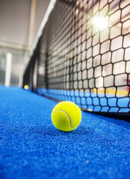 A bright yellow padel ball sitting on the blue synthetic turf of a court against a net.