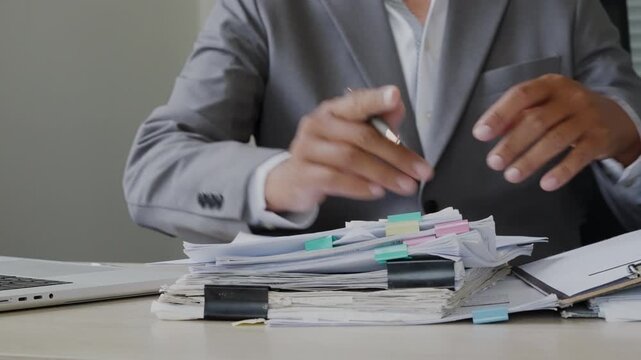Business executive writing and organizing paper files beside a laptop, showing document control, audit preparation, contract approval, financial reporting, and office workload.