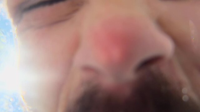 Bearded man plunging face into water and shaking head above underwater camera, exhaling bubbles, wiping wet skin, blue sky, sun flare and leaves in extreme low angle authentic outdoor freshness shot