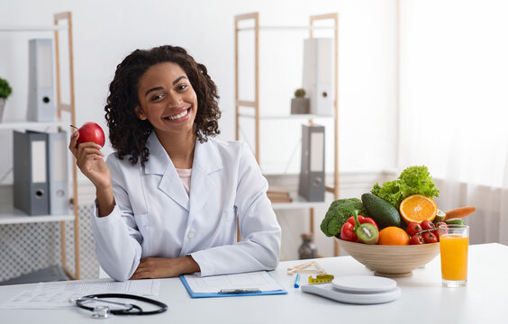Diet while lockdown. Pretty black lady dietologist holding apple in her hand and smiling, recommending fresh fruits, posing at clinic, copy space