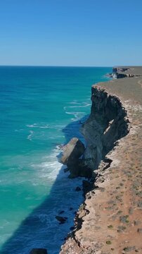 Vertical drone shot pulling back above the Bunda Cliffs, following the coastline with blue sky and ocean stretching into the distance