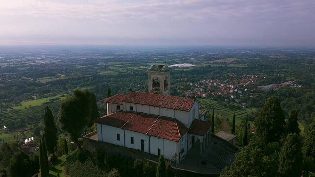Sanctuary of Beata Vergine del Carmelo on hilltop surrounded by cypress trees and vast landscape in Montevecchia, Italy. Aerial backward ascending, copy space