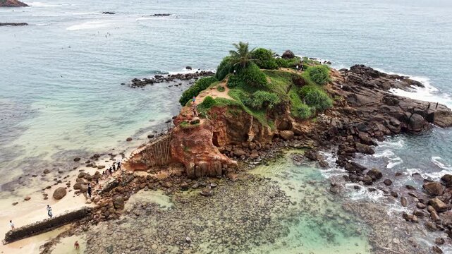 Aerial view of Parrot Rock islet with people walking along the narrow sandbar surrounded by turquoise ocean waves in Mirissa, Southern Province, Sri Lanka.