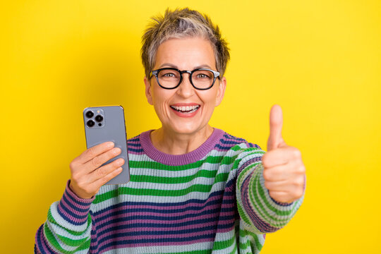 Joyful mature woman with glasses holds smartphone and gives thumbs up against a bright yellow background