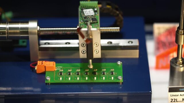 A high-precision linear actuator moves a testing probe over an electronic circuit board during a technical quality control demonstration.