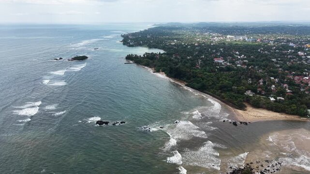 Aerial view of the tropical coastline with waves crashing against rocks and lush green trees along the sandy beach in Matara, Southern Province, Sri Lanka.