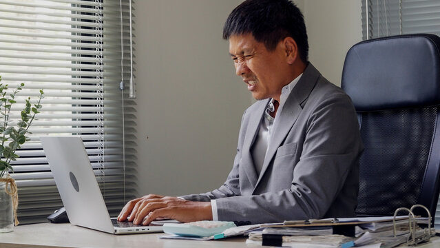 Man sits focused at laptop in office. Hands rest on keyboard, eyes concentrated. Light filters through blinds behind him. Desk holds papers, pen, and small plant