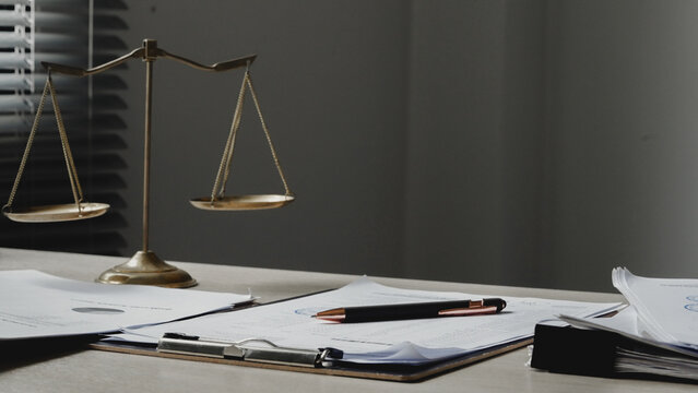 Golden scales rest on a cluttered desk. Papers, pens, and folders suggest legal work. Soft light highlights the courtroom symbolism. Serious tone conveys legal responsibility. Ideal for legal, justice