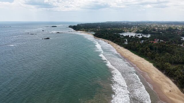 Aerial view of the scenic tropical coastline featuring a sandy beach, turquoise ocean waves, and dense palm tree forests under a cloudy sky in Matara, Southern Province, Sri Lanka.