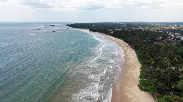 Aerial view of the turquoise ocean waves crashing on a sandy beach lined with palm trees and coastal buildings in Matara, Southern Province, Sri Lanka.