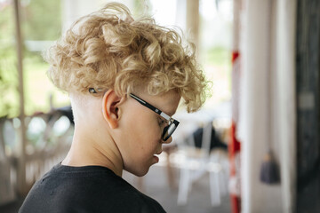 Young boy with curly blonde hair wearing black t-shirt and glasses stands outdoors, facing away from the camera with blurred background of chairs and greenery © Jolanta
