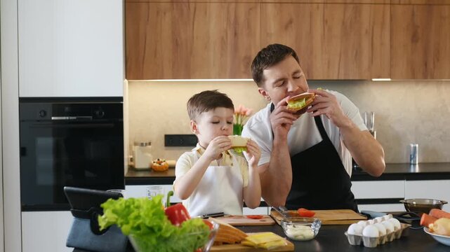 Father and son enjoy making sandwiches together in a contemporary kitchen, surrounded by fresh ingredients and kitchenware on the countertop