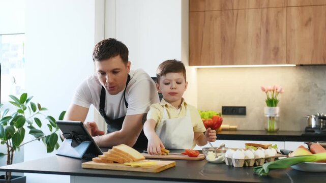 Father and son engage with a tablet in a contemporary kitchen, surrounded by food ingredients including bread, ham, and tomato on the countertop