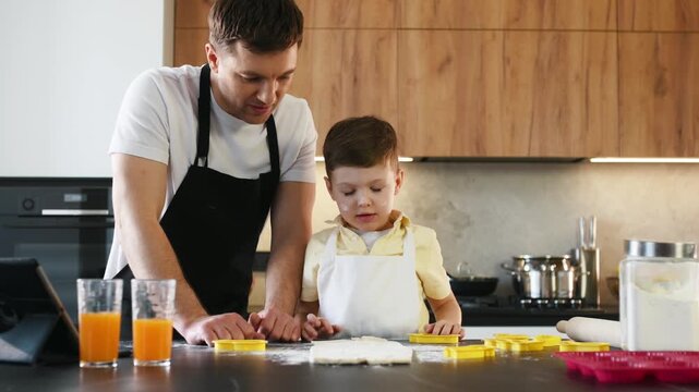 Father and young son baking together in a modern kitchen, using cookie cutters on flour-covered countertop with orange juice glasses and kitchen tools nearby